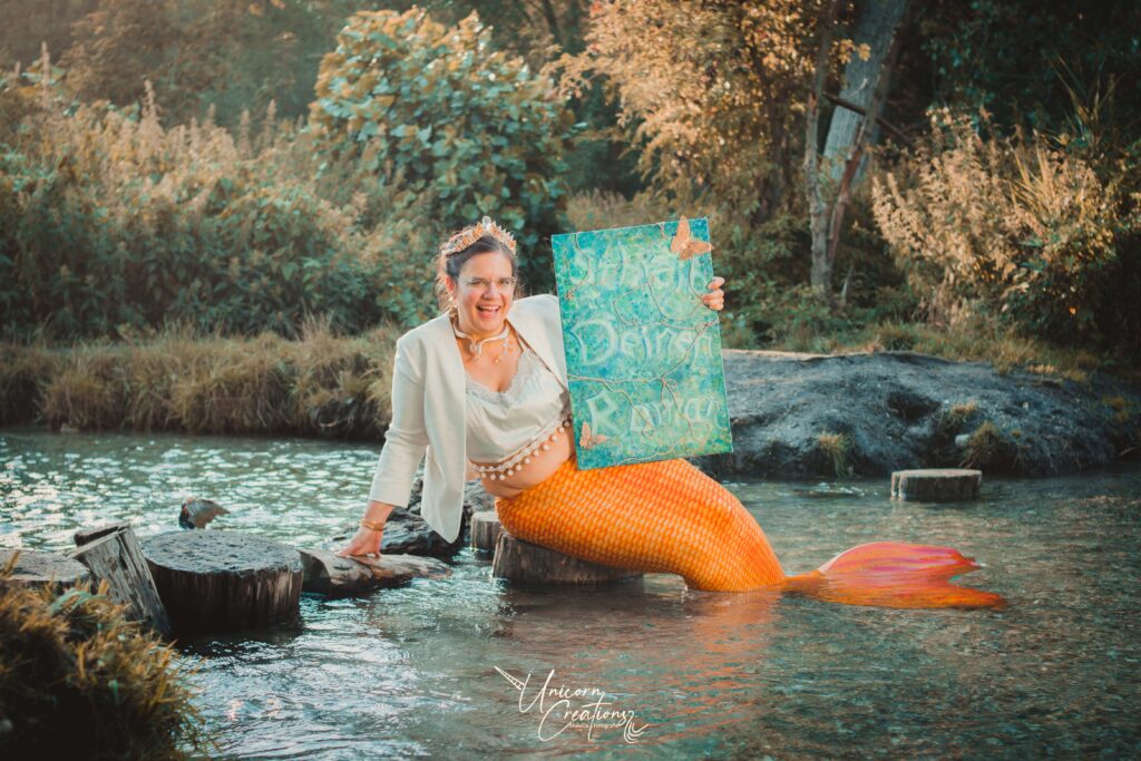 Hanna als Seejungfrau sitzt im Wasser und lächelt strahlend in die Kamera. Sie hält ein selbstbemaltes Schild: Schreib Deinen Roman.