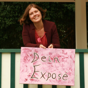 Bettina lächelt verträumt hinter einem Geländer. Sie trägt einen dunkelroten Blazer hält ein farblich passendes Schild mit der Aufschrift "Das Exposé".