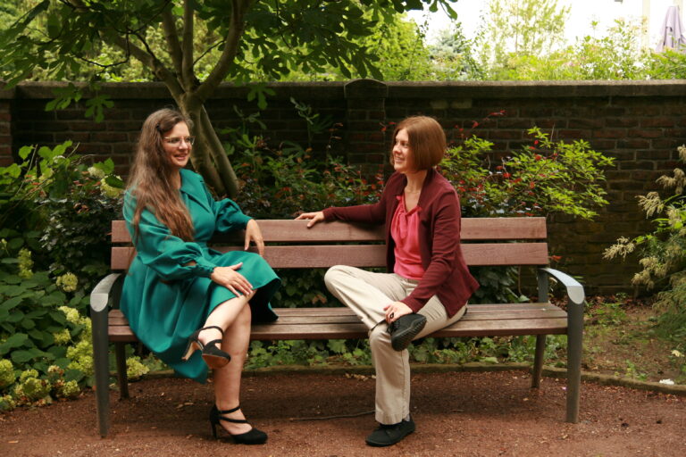 Hanna und Bettina sitzen auf einer Bank, hinter der sich eine Mauer, Büsche und ein schiefgewachsener Baum befinden. Sie scheinen in ein angeregtes Gespräch verwickelt zu sein.