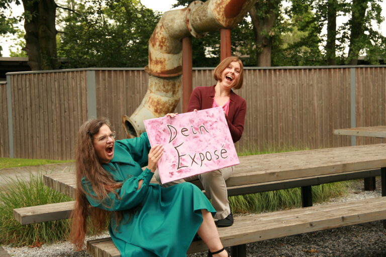 Hanna und Bettina sitzen auf einer Bank und einem Tisch. Sie zerren aus zwei Richtungen an einem Schild "Das Exposé" und haben offenkundig Spaß dabei.